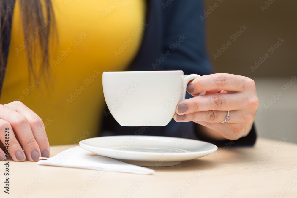 hand with painted rings with a ring holding and lifting a small white cup, there is a plate and napkin, on a smooth wooden surface, in the background you can see the yellow shirt of the person holding hand with painted rings with a ring holding and lifting a small white cup, there is a plate and napkin, on a smooth wooden surface, in the background you can see the yellow shirt of the person holding