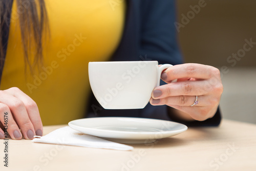 hand with painted rings with a ring holding and lifting a small white cup  there is a plate and napkin  on a smooth wooden surface  in the background you can see the yellow shirt of the person holding