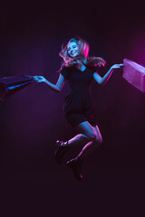 Jumping, flying high with shopping bags, laughting. Portrait of young woman in neon light on dark backgound. The human emotions, black friday, cyber monday, purchases, sales, finance concept.