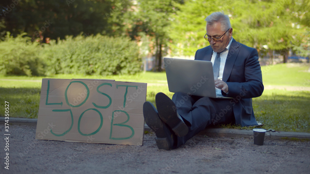 Businessman sitting on street with lost job cardboard sign and paper cup for charity and sing laptop Businessman sitting on street with lost job cardboard sign and paper cup for charity and sing laptop