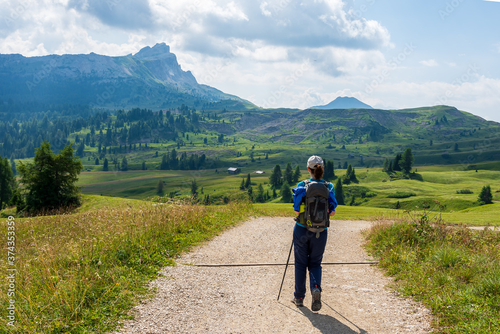 Young woman with a hat, trekking poles, a backpack hiking on a trail through green meadows in the Italian Alps. Dolomite peaks are visible in the background. Val Badia, South Tyrol - Italy