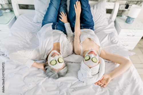 Top angle view of two charming women, senior gray haired lady and young girl with towel on head, lying on the bed and enjoying spa day with face masks and cucumber slices on face