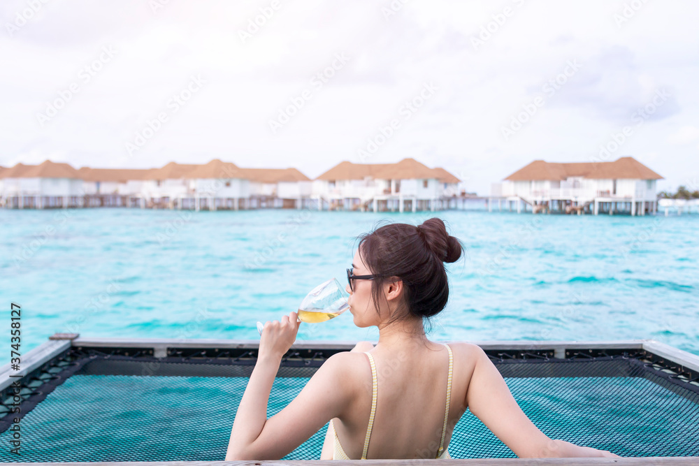 Fototapeta premium Young beautiful girl in a hammock with wine glass over the blue sea. Young girl enjoying glass of wine by the sea. Young woman watching sunset from sea hammock enjoying tropical vacations.