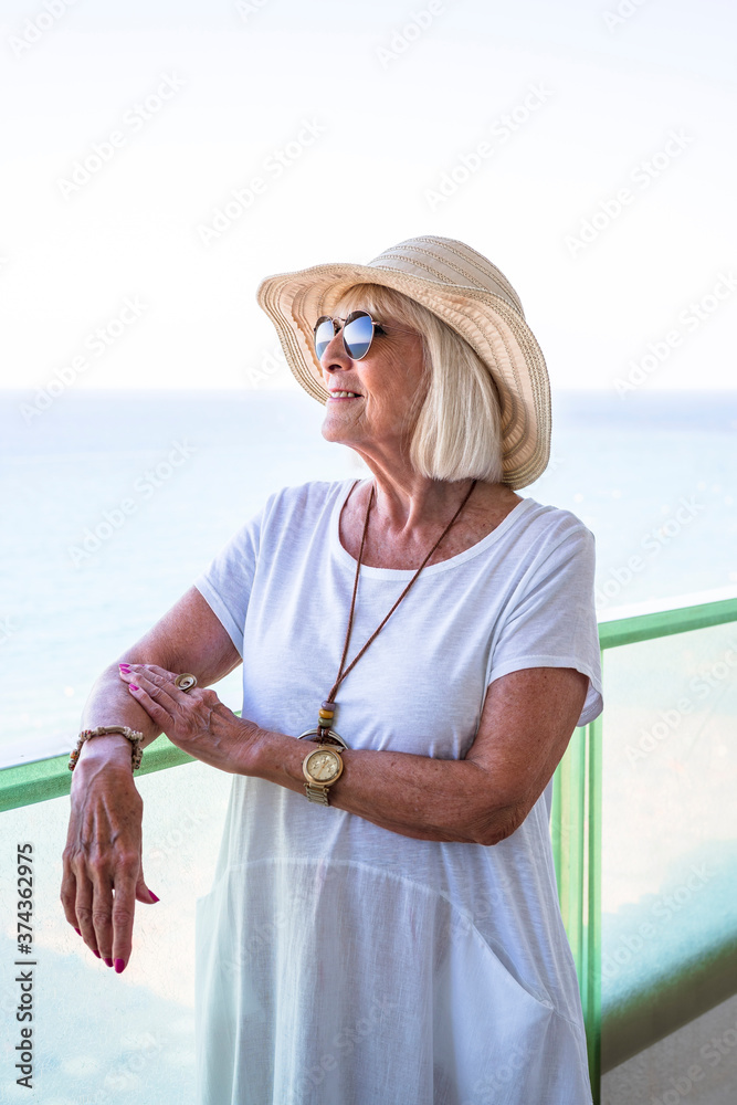 Senior woman leaning on a terrace railing looking out to sea wearing a straw hat and white dress