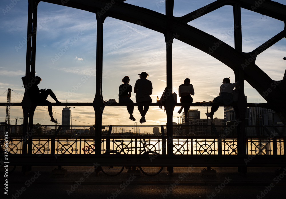 Fototapeta premium Unrecognizable backlit people enjoying the sunset sitting on a steel bridge construction over railway tracks called Hackerbrücke. Munich, Germany.
