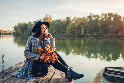 Fotografie Woman relaxing on lake pier by boat admiring autumn landscape holding branches with leaves