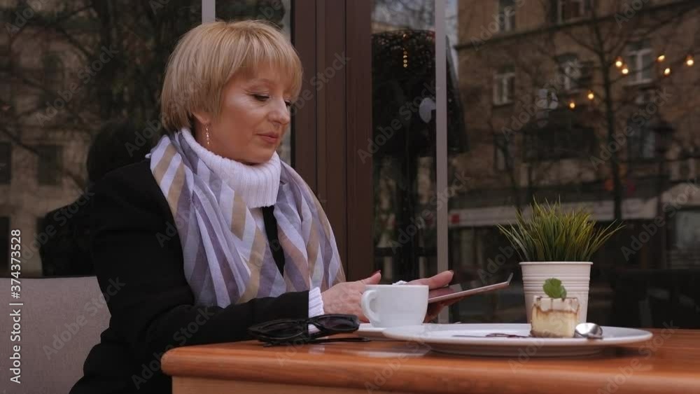 An elderly woman is relaxing in a street cafe, she uses a tablet and drinks coffee. An elderly woman is relaxing in a street cafe, she uses a tablet and drinks coffee.