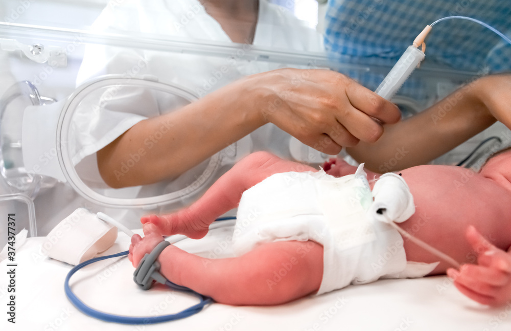 Photo of a premature baby in incubator. Focus is on his feet. Nurse is