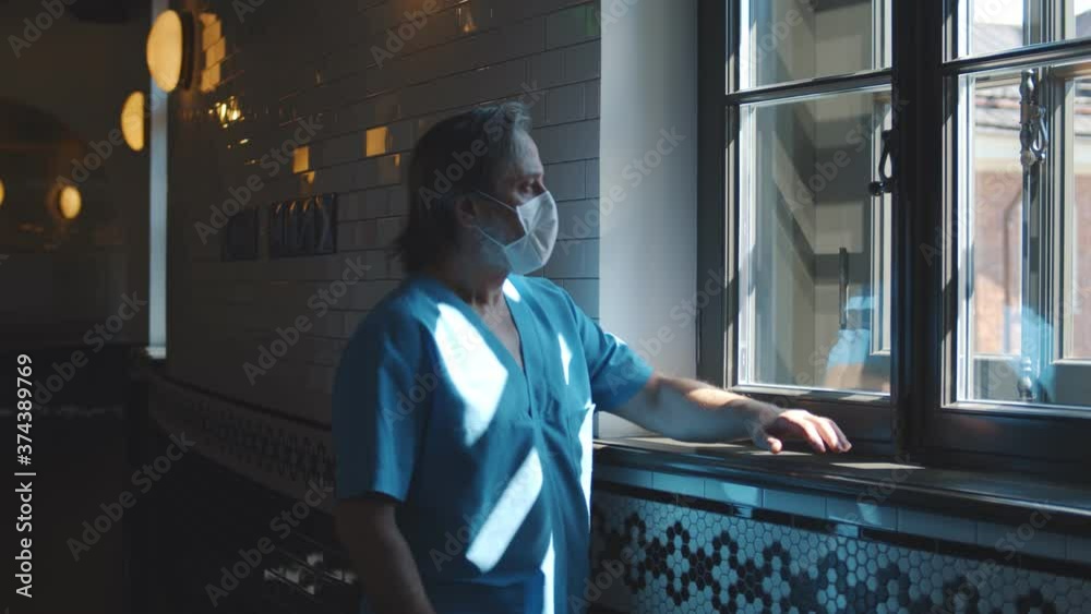 Senior doctor in uniform and medical mask resting looking through window in hospital corridor Senior doctor in uniform and medical mask resting looking through window in hospital corridor
