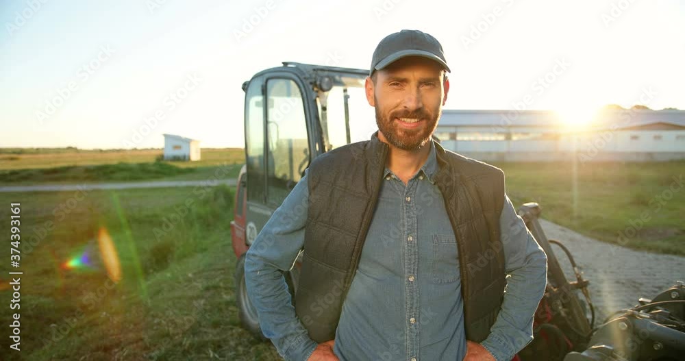 Portrait of young Caucasian handsome happy man farmer standing in field and smiling to camera. Big tractor on background. Cheerful male worker in agricultural farm. Sunlight. Agriculture farming.