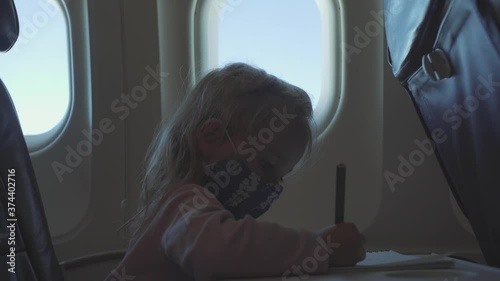 Child, during the COVID-19 pandemic, flies on an airplane wearing a mask and draws in an album