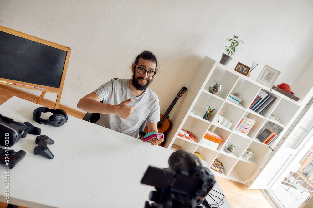 Male technology blogger in glasses showing thumbs up, holding game controller joystick while recording video blog or vlog about new gadgets at home