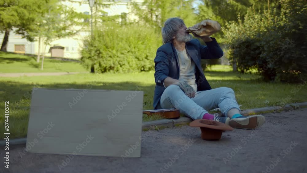 Senior homeless man sitting on ground and drinking alcohol with blank cardboard sign Senior homeless man sitting on ground and drinking alcohol with blank cardboard sign