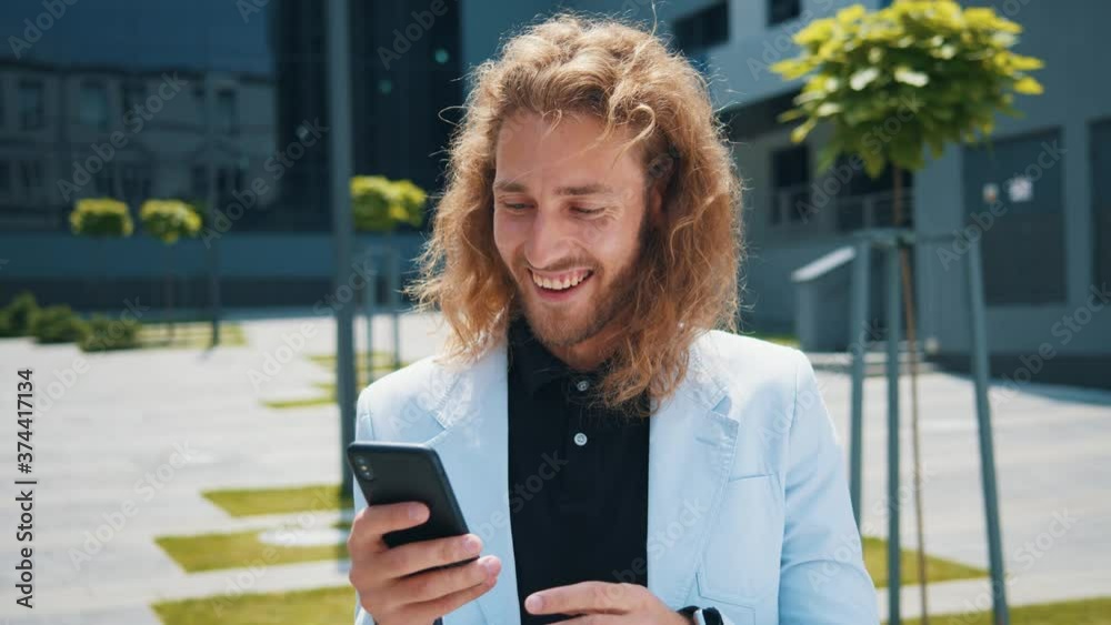 Euphoric ypoung entrepreneur woith long hair using smartphone clenching fist overjoying victory reading fantastic business news online. Success. Achievement concept.