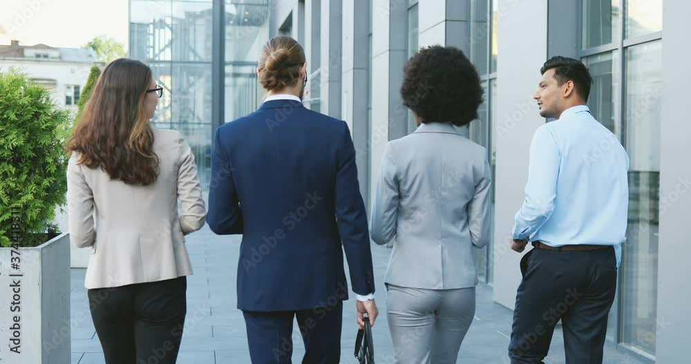 Rear of mixed-races young people in business style walking the street and talking. Multiethnic business white-collars males and females going to work in morning and chatting. Men and women. Back view.