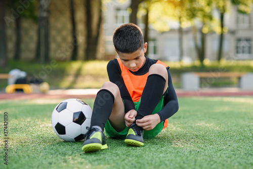 Cute 13-aged football player on artificial green covering of outdoors sport field and tying the shoelace on his boots. © gorynvd