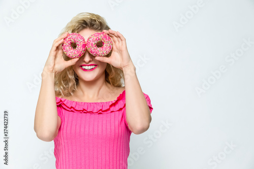 Food Ideas. Portrait of Happily Smiling  Caucasian Blond Holding Pink Doughnuts in Both Hands in Front of Her Face. Posing On White.