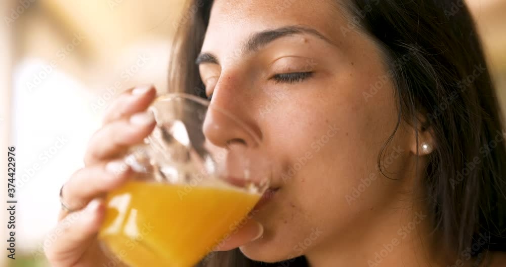 Authentic close up shot of an young brunette woman is drinking a fresh healthy dietetic biological natural orange juice from a transparent glass  taking a breakfast in the morning.