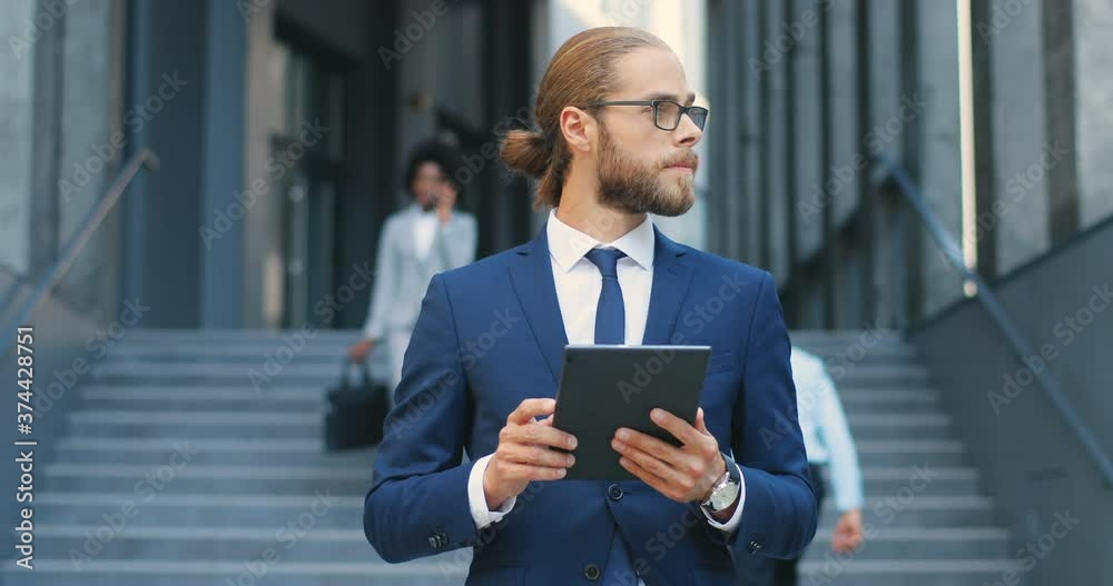 Handsome Caucasian young businessman in glasses, suit and tie using tablet device at street. Man in business style tapping and scrolling on gadget computer. Outdoor. At business center building. Handsome Caucasian young businessman in glasses, suit and tie using tablet device at street. Man in business style tapping and scrolling on gadget computer. Outdoor. At business center building.
