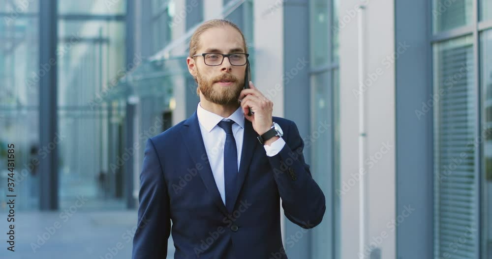 Handsome young Caucasian man in glasses walking and talking on mobile phone in city center at modern building. Male speaking on cellphone and strolling. Stylish rich businessman. Telephone call. Handsome young Caucasian man in glasses walking and talking on mobile phone in city center at modern building. Male speaking on cellphone and strolling. Stylish rich businessman. Telephone call.