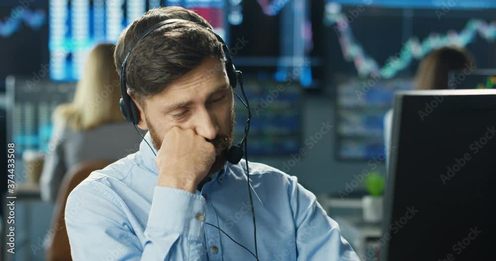 Young handsome Caucasian bored man broker in headset working at computer monitor and yawning. Boring workday. Male trader listening to client in call center. Young handsome Caucasian bored man broker in headset working at computer monitor and yawning. Boring workday. Male trader listening to client in call center.
