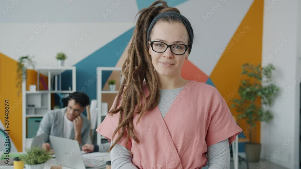 Portrait of cheerful young woman with dreadlocks and piercing standing in office smiling looking at camera while people are working in background Portrait of cheerful young woman with dreadlocks and piercing standing in office smiling looking at camera while people are working in background