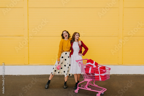 Full length view of two ladies standing near yellow wall. Girls posing after shopping.