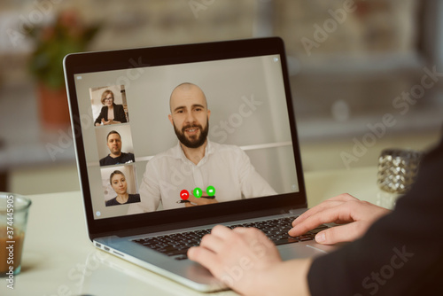 A laptop screen view over a woman's shoulder. A girl is listening to a statement of her colleague on an online briefing on a computer in her studio. A student on an online video meeting on a computer.