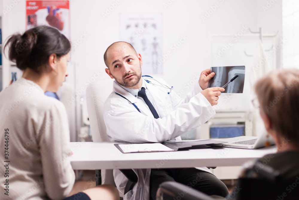 Doctor showing knee injury on x-ray image in hospital office to young woman and her disabled mother in wheelchair. Medic wearing white coat and stethoscope.
