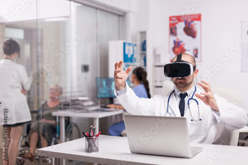 Doctor using futuristic augmented reality technology wearing headset in hospital office and white coat with stethoscope. Disabled senior woman discussing with medic on clinic corridor.