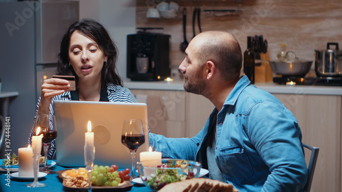 Modern happy couple shopping online on laptop during romantic dinner. Adults sitting at the table, searching, browsing, surfing, using technology card payment, internet