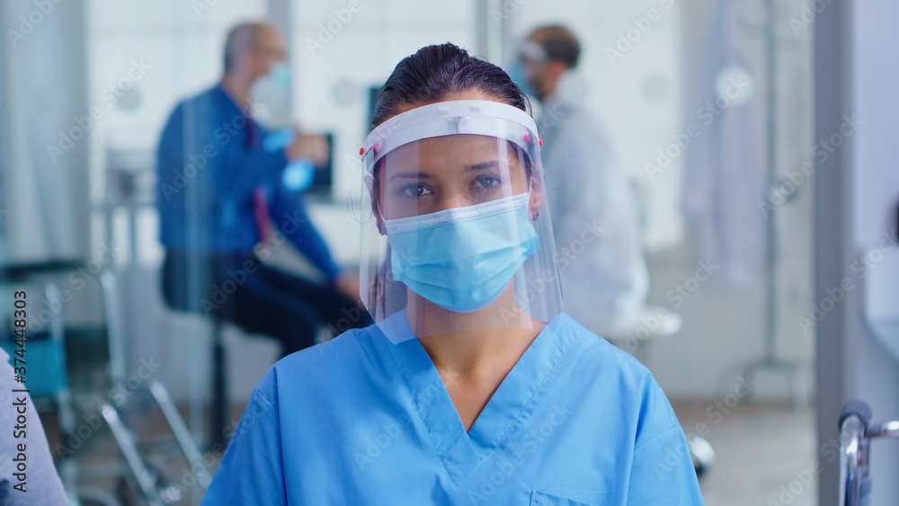 Medical assistant with visor and face mask against coronavirus looking at camera in hospital waiting area. Doctor consulting senior man in examination room. - obrazy, fototapety, plakaty 
