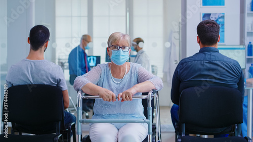 Disabled senior woman with walking frame in hospital waiting area looking at camera. Mature woman wearing face mask against coronavirus. Nurse in examination room.