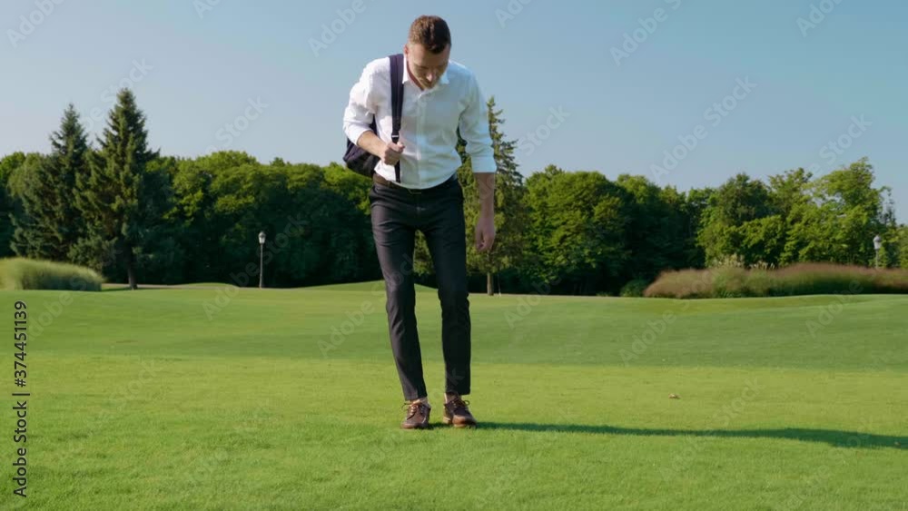 Young businessman with backpack on shoulder walks in the park on the green grass. Man in classic white shirt and trousers sits on the grass. 4K slow motion footage Young businessman with backpack on shoulder walks in the park on the green grass. Man in classic white shirt and trousers sits on the grass. 4K slow motion footage