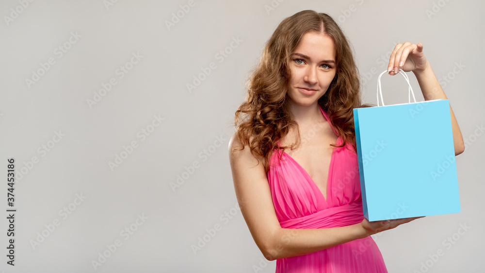Shopping assistant. Special offer. Elegant woman in pink dress showing brand mockup blue paper bag isolated on light gray copy space background. Personal stylist. Sale discount. Shopping assistant. Special offer. Elegant woman in pink dress showing brand mockup blue paper bag isolated on light gray copy space background. Personal stylist. Sale discount.