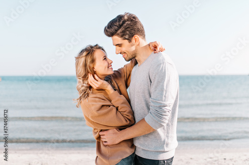 Graceful blonde girl looking at boyfriend with love on nature background. Outdoor photo of happy man dancing with wife beside sea.