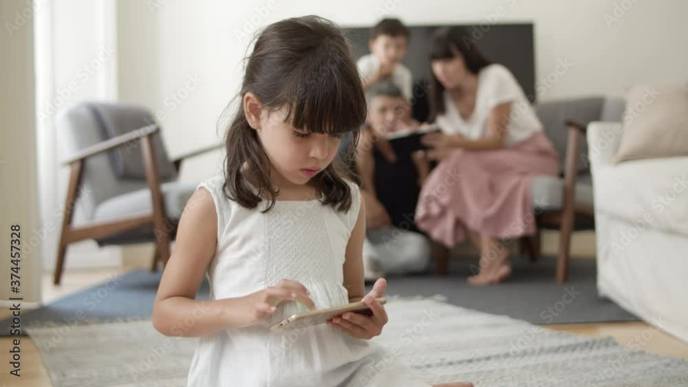 Young girl smiling at camera, then using smartphone for browsing internet while her parents and brother using digital device in background. Communication or internet concept Young girl smiling at camera, then using smartphone for browsing internet while her parents and brother using digital device in background. Communication or internet concept