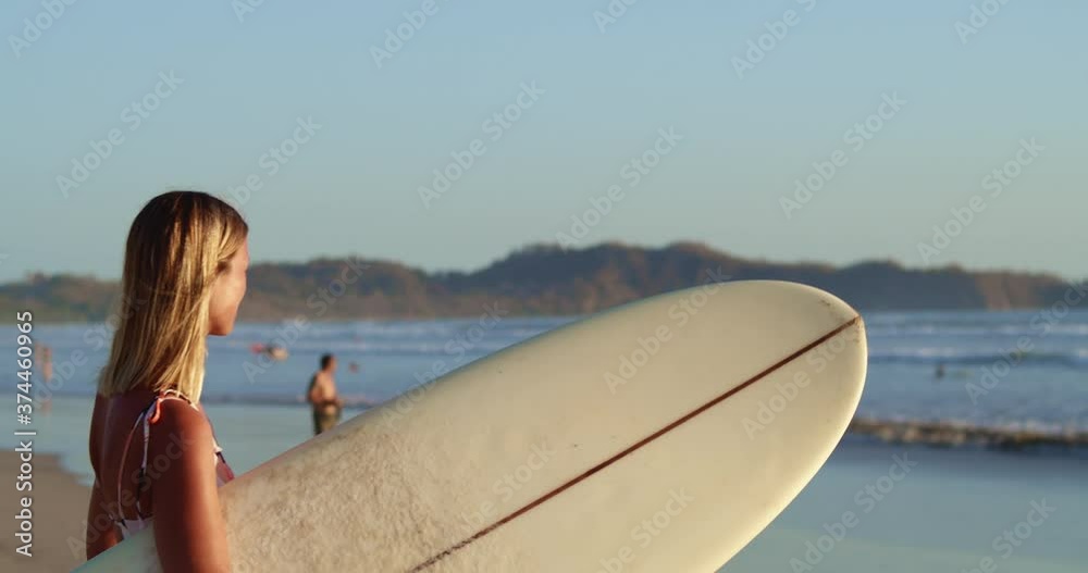 Young blonde and tanned surfer looks out at ocean as sun sets in the distance seeing where the best waves are to surf