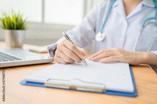 healthcare and medical care working from home concept, beautiful female Asian doctor working in home office smiling writing one clipboard diagnosing patients health using computer laptop and tablet