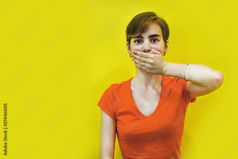 A young woman with short hair covered her mouth with her hands. Isolated on a yellow background. Keep it secret. space for text. Concept: silence, no talking, I won't say anything. Selective focus A young woman with short hair covered her mouth with her hands. Isolated on a yellow background. Keep it secret. space for text. Concept: silence, no talking, I won't say anything. Selective focus