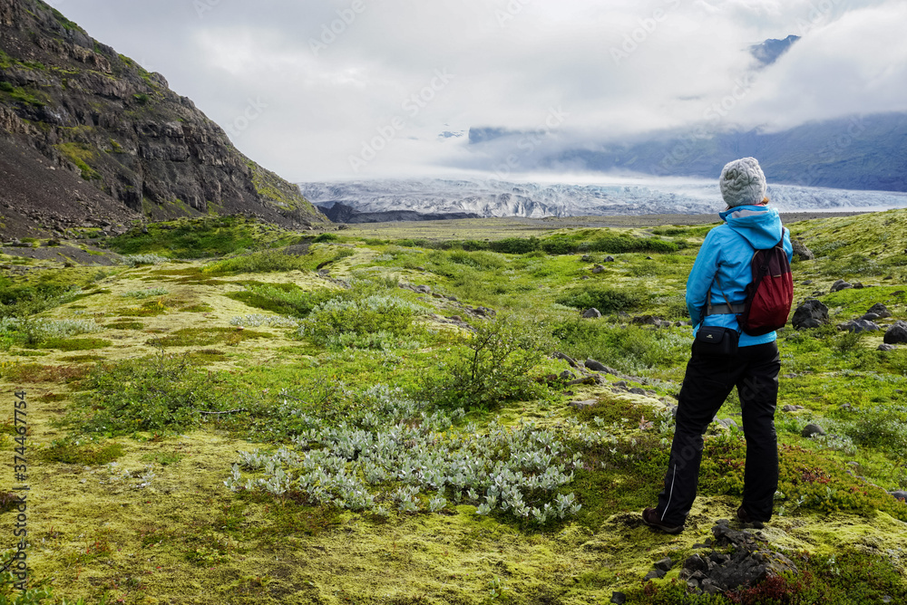mountains, a green field and a glacier in thick clouds, a girl in sports clothes look towards the glacier, nature of Iceland