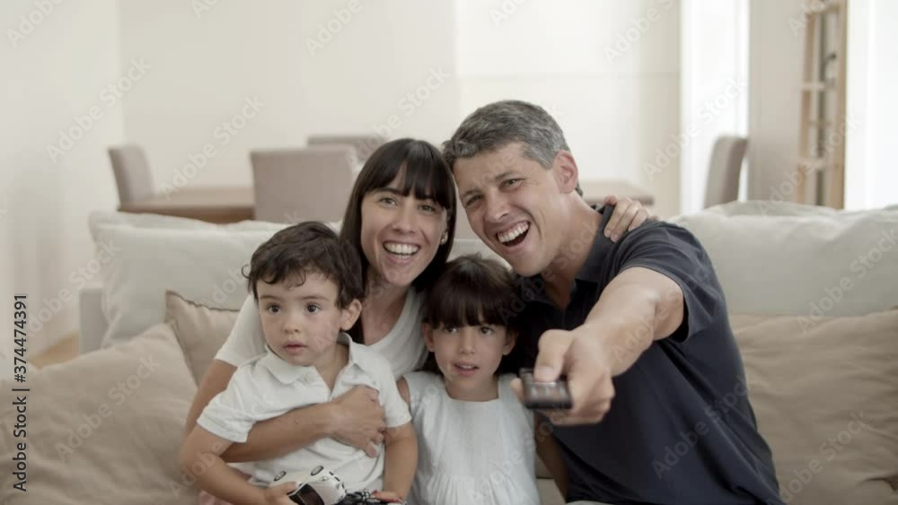 Happy parents couple with two kids watching favorite show on TV, sitting o couch, pointing remote control at camera. Front view. Television concept Happy parents couple with two kids watching favorite show on TV, sitting o couch, pointing remote control at camera. Front view. Television concept