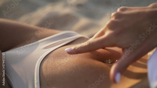 A tanned sexy slim girl in a white underwear with a piercing in the navel lies on the sand on the beach. Hand touching belly in slow motion