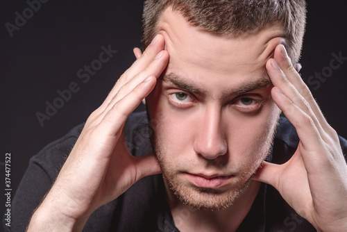 Young handsome man in a black shirt sits and holds his head on a black background. Experiencing stress, pain