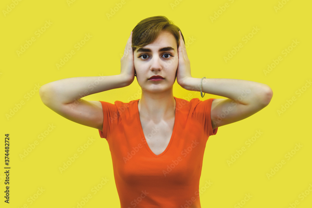 A young woman with short hair holds her head with her hands. Calm expression, big eyes, covers his ears with his hands. Isolated on a yellow background. Concept: fright, headache, hear nothing. A young woman with short hair holds her head with her hands. Calm expression, big eyes, covers his ears with his hands. Isolated on a yellow background. Concept: fright, headache, hear nothing.