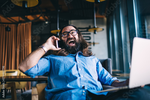Joyful hipster male with laptop talking on cellphone in cafe