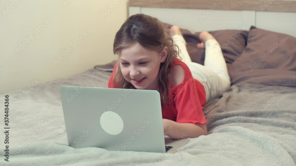 Young beautiful brunette girl lying on the bed at home communicates with relatives online. There is a laptop on the bed. Young beautiful brunette girl lying on the bed at home communicates with relatives online. There is a laptop on the bed.