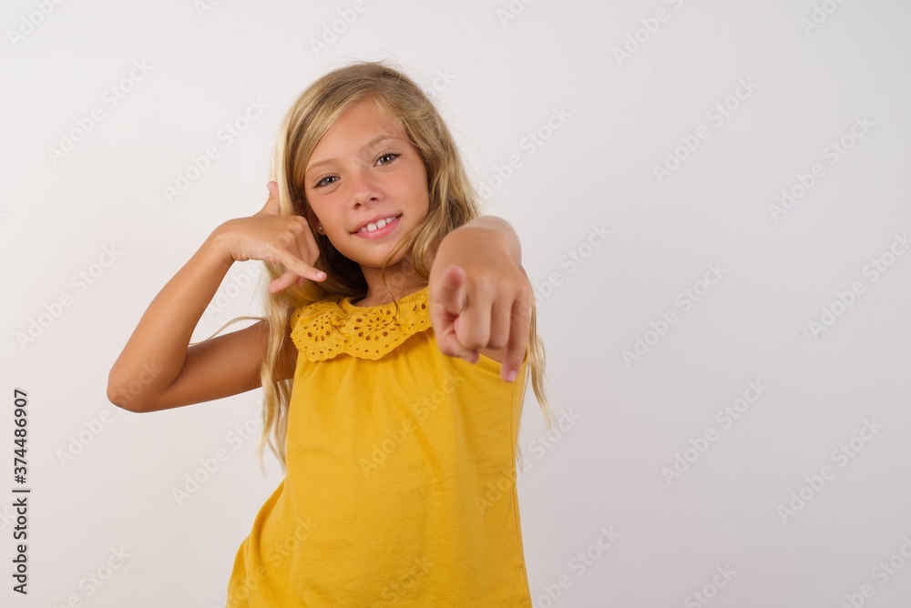 Little blond girl wearing yellow dress over white background smiling cheerfully and pointing to camera while making a call you later gesture, talking on phone Little blond girl wearing yellow dress over white background smiling cheerfully and pointing to camera while making a call you later gesture, talking on phone