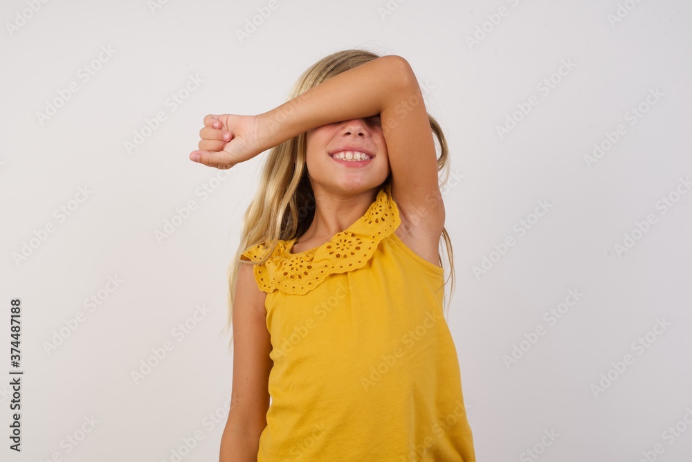 Little blond girl wearing yellow dress over white background covering eyes with arm smiling cheerful and funny. Blind concept. Little blond girl wearing yellow dress over white background covering eyes with arm smiling cheerful and funny. Blind concept.