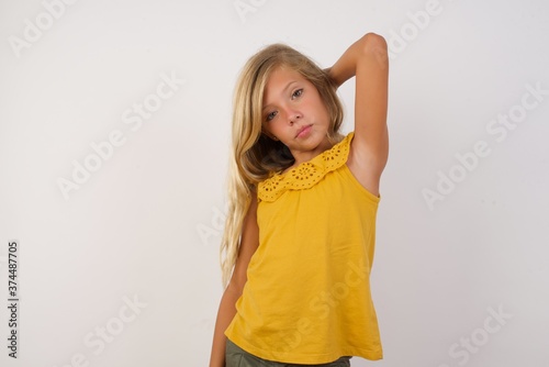 Young blonde kid girl wearing yellow dress over white background confused and wonders about something. Holding hand on her head, uncertain with doubt. Pensive concept.